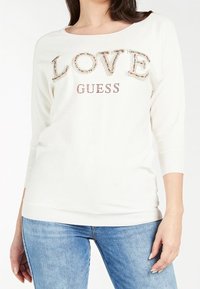 Maglia bianca a maniche lunghe con la scritta "LOVE" in decorazioni colorate e "GUESS" in strass. Silhouette aderente e polsini a costine.