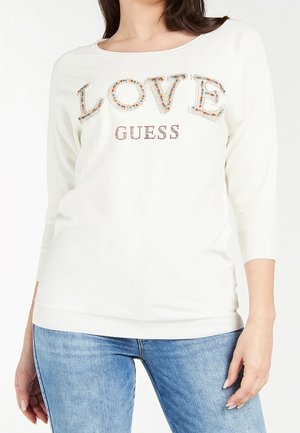 Haut à manches longues blanc avec "LOVE" en embellissements colorés et "GUESS" en strass. Silhouette ajustée et poignets côtelés.