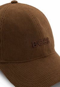 Brown corduroy cap featuring a ribbed texture and an embroidered logo on the front. The design includes a curved brim and top button detail.
