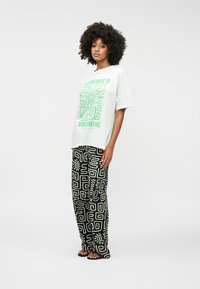 T-shirt blanc graphique avec le texte vert "SUMMER DREAMING" associé à un pantalon noir à motifs comportant des designs abstraits blancs. Chaussures décontractées.