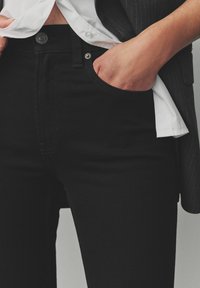 Jeans de mezclilla negra con un diseño ajustado, que cuentan con un cierre de botón, bolsillos delanteros y una ligera textura. Combinados con una camisa blanca.