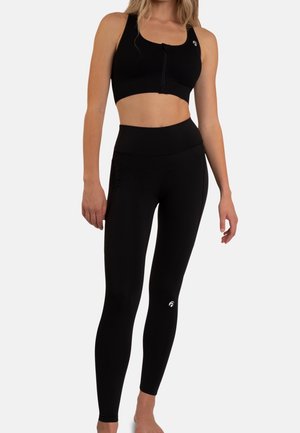 Kvinde iført sort sports-BH med lynlås foran og højtsiddende sorte leggings med små hvide logoer, stående mod en hvid baggrund.