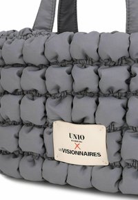 Bolso tote gris acolchado con un diseño de colcha, con una etiqueta blanca que dice "UNIO HAMBURG X LES VISIONNAIRES." La textura de la tela es suave y lisa.