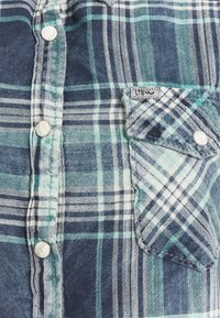 Gros plan d’une chemise à carreaux bleue et verte avec des boutons blancs et une petite poche sur le côté droit comportant une étiquette en tissu.