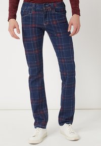 Jeans de cuadros en azul marino que presentan un patrón de líneas rojas y blancas, cinco bolsillos y un corte recto, combinados con zapatillas blancas.