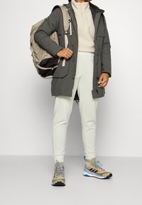 Szary parka, beżowa bluza polarowa, białe joggery i brązowo-niebieskie sneakersy. Plecak ma elegancki design z wieloma przegrodami i regulowanymi paskami.