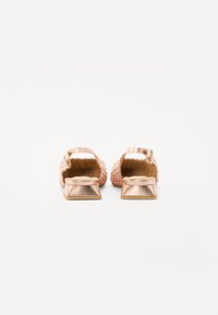 Sandalias de tacón bloque en oro rosa con parte superior trenzada, espalda abierta y correa con hebilla. La textura es suave con un acabado metálico y suela plana.