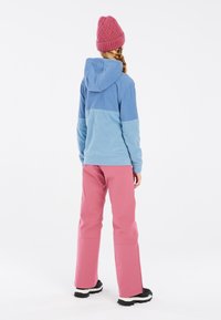 Veste en polaire bleue avec capuche, accent rose, assortie à un pantalon rose et des bottes noires. Design simple et fonctionnel avec un look en couches.