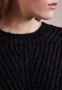 Das Untergesicht und der Hals einer Frau sind sichtbar, die einen dicken, schwarzen, gerippten Strickpullover trägt, mit leicht zur Seite gedrehtem Kopf.