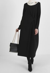 Vestido negro de manga larga con un detalle de nudo retorcido en la cintura. Combinado con un bolso negro texturizado con cadenas en las asas y una bufanda blanca.