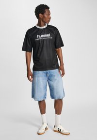 Black short-sleeve jersey with a ribbed texture, featuring white “hummel PERFORMANCE” branding, paired with blue denim shorts and white sneakers.