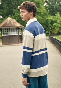 Long-sleeve polo shirt in blue and beige with horizontal stripes. Soft fabric with a collar and relaxed fit, paired with blue denim jeans.
