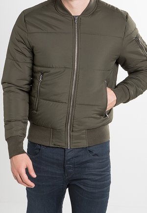 Homme portant une veste bomber matelassée vert olive avec poches zippées, une main dans la poche, associée à un jean bleu foncé.