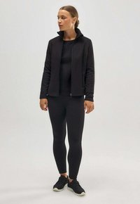 Chaqueta deportiva negra con collar alto, cierre de cremallera, combinada con una camiseta negra ajustada y leggings. El modelo lleva zapatillas negras. Textura suave.