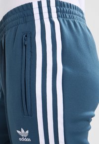 Teal träningsbyxor med vit trippelrand-design, elastisk midja, sidoficka med dragkedja och broderad logotyp på nedre benet.