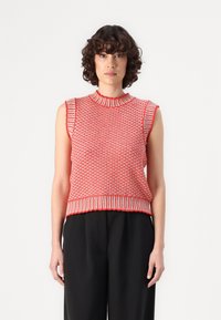 Rotes gestricktes ärmelloses Oberteil mit einem strukturierten Muster, geripptem Ausschnitt und Saum mit kontrastierenden Akzenten. Kombiniert mit schwarzen Hosen.