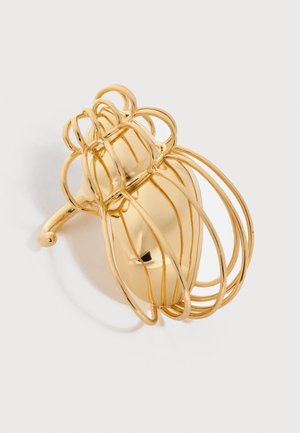 Agrafă de păr din metal cu nuanță aurie, în formă de clește, cu un design curbat de sârmă deschisă și centru neted și solid, proiectând o umbră pe un fundal deschis.