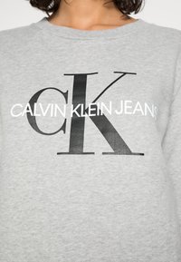 Sweatshirt gris avec un grand logo noir "CK" et le texte "CALVIN KLEIN JEANS" en blanc, doté d'une texture douce et d'un design classique à col rond.