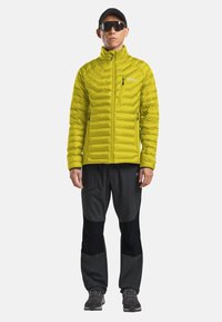 Giacca isolante verde lime con cuciture a zigzag, tasche con zipper, abbinata a pantaloni softshell neri e scarpe da escursionismo grigie.