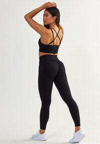 Czarny stanik sportowy z krzyżującymi się ramiączkami, w połączeniu z czarnymi legginsami z wysokim stanem. Ubranie ma gładką fakturę i dopasowany krój.