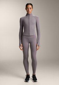 Svetlofialový športový set zahŕňajúci zipsovací dlhý rukáv a priliehajúce legíny, vyrobený z hladkého, elastického materiálu. Súčasťou sú čierne tenisky.