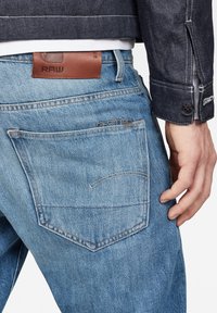 Jeans de mezclilla azul claro con corte recto, con bolsillos traseros y una etiqueta de cuero marrón que muestra "RAW". La textura está ligeramente descolorida.