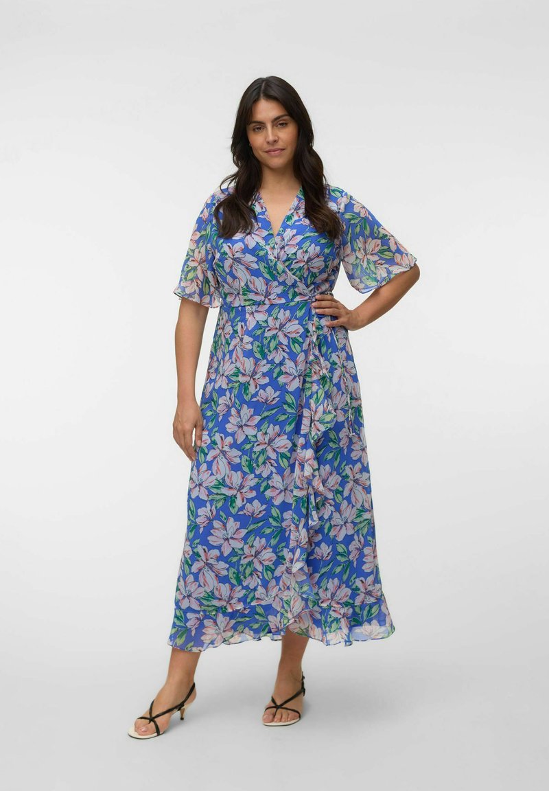 Femme portant une robe portefeuille bleue à fleurs avec des manches courtes transparentes, debout avec une main sur la taille et portant des talons noirs à brides.