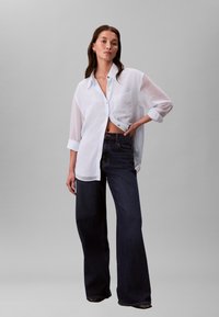 Chemise blanche boutonnée avec une poche poitrine, associée à un jean denim bleu foncé à jambes larges. Tissu lisse avec manches retroussées, coupe décontractée.