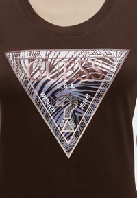 Καφέ μπλούζα με μεγάλο γυαλιστερό τριγωνικό λογότυπο Guess με σχέδιο ζέβρας, ερωτηματικό και τις λέξεις "U.S.A. Washed Jeans."