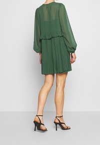 Vestido verde plisado con mangas largas y transparentes, una cintura con volantes y una silueta fluida. Sandalias negras de tacón alto completan el conjunto.