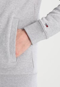 Grijze sweatshirt met een kangoeroe-pocket, ribgebreide mouwen en een klein merkdetail met rood, wit en blauwe strepen op de mouw.
