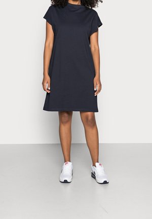 Femme portant une robe bleu marine à manches courtes et longueur au genou, associée à des baskets blanches, se tenant devant un fond gris clair uni.