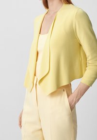 Keltainen lyhyt neulecardigan pehmeästä langasta avonaisella edellä. Pia keltainen toppi ja muotoillut housut.