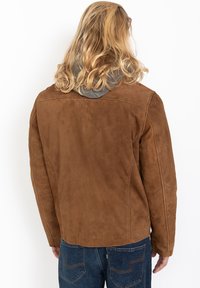 Persona con lunghi capelli biondi che indossa una giacca in suede marrone e jeans blu, di spalle su uno sfondo bianco semplice.