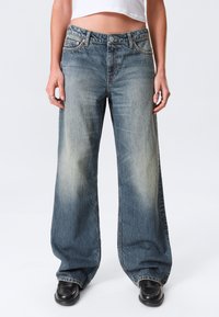 Μπλε τζιν παντελόνια με φαρδιά λουρίδα, ξεθωριασμένο πλύσιμο, σχεδιασμό με πέντε τσέπες, πορτοκαλί ραφές και κλείσιμο με κουμπιά. Λείο υλικό, άνετη εφαρμογή.