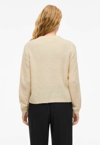 Pull en mailles couleur crème avec une finition texturée, col rond et manches longues. Ourlet légèrement effiloché, associé à un pantalon noir.