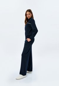 Marinblå träningsdräkt med en jacka med blixtlås och vit kant, elastisk midja och vida byxor. Stylad med vita sneakers mot en ljus bakgrund.