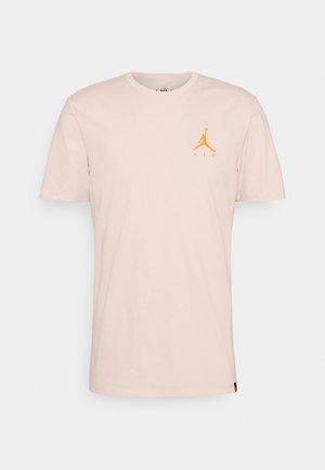 Lyse beige T-shirt med korte ærmer med lille orange Jumpman-logo og "AIR" tekst på venstre bryst, vist på ensfarvet baggrund.