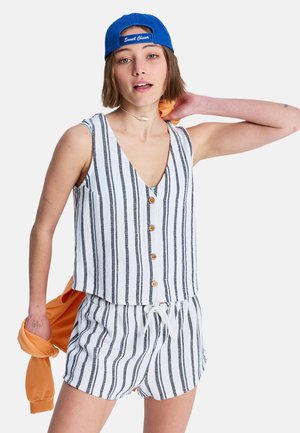 Junge Frau in ärmellosem weiß-schwarz gestreiftem, geknöpftem Oberteil und passenden Shorts, trägt eine blaue Kappe und hält eine orangefarbene Jacke.