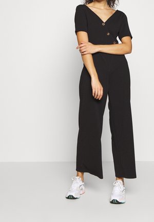 Kobieta ubrana w czarny kombinezon z krótkim rękawem i guzikami oraz białe trampki, stojąca z założonymi ramionami na jednolitym tle.