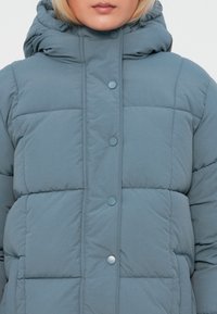 Sieviete, kas valkā gaiši zilu quiltētu pūdera jaku ar augstu apkakli un spiedpogu aizdari, īsu līdz zem deguna uz baltas fona.