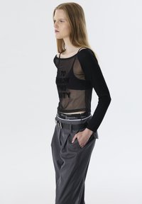 Zwarte lange mouwen mesh top met de tekst "DIT FEEST GOOIEN", gecombineerd met hoog getailleerde grijze geplooide broek en een zwarte riem met hardwaredetails.