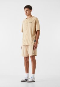Chemise beige en maille à manches courtes avec des détails de boutons, accompagnée de shorts assortis, le tout présentant un branding minimal et une coupe décontractée.