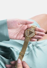 Guldring med mesh-rem, der har et rundt urens ansigt pyntet med klare krystaller og et trekantet designelement. Holdes i en hånd.