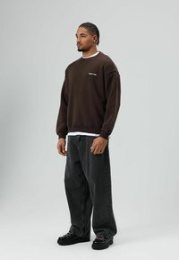 Bruine trui met een losse pasvorm, witte gelaagde blouse eronder, gecombineerd met losse zwarte jeans en zwarte sneakers met kleurrijke veters.