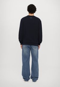 Sweatshirt azul marinho de mangas longas com punhos e bainha canelados. Apresenta uma pequena etiqueta de logo na parte de trás. Combinado com jeans de perna larga em azul claro.