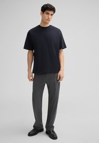 Zwarte T-shirt met korte mouwen van zachte stof, gecombineerd met grijze broek met streepjes en zwarte instappers, staand tegen een lichtgrijze achtergrond.
