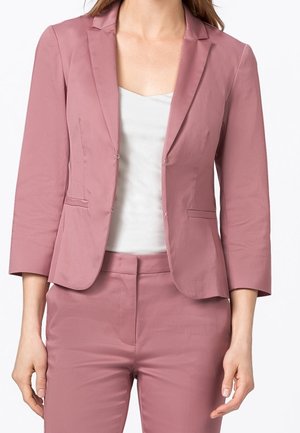 Blazer rose ajusté avec des manches trois-quarts, fermeture à un bouton et deux poches avant, assorti à un haut blanc et un pantalon coordonné.