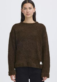 Maglione marrone, texture morbida, con vestibilità ampia, collo rotondo e spalle scese. Presenta un'etichetta sul bordo e maniche lunghe ripiegate.