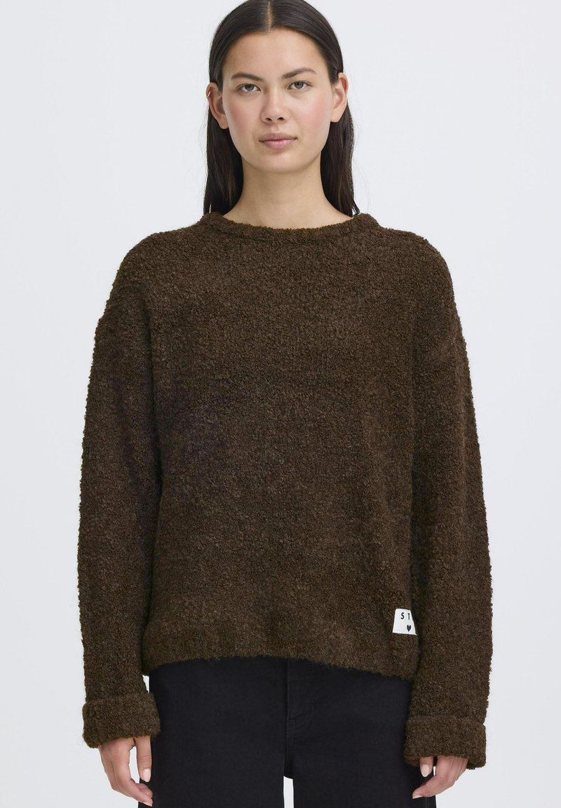 Maglione marrone, texture morbida, con vestibilità ampia, collo rotondo e spalle scese. Presenta un'etichetta sul bordo e maniche lunghe ripiegate.